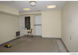 This is a photograph of an empty commercial interior space in the process of renovation or construction. The room features cream-colored walls, fluorescent ceiling fixtures, and concrete flooring with protective tape marking borders. A single wooden 
