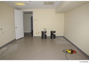 This photograph documents an empty interior room with cream-colored walls, recessed ceiling lights, and concrete flooring. The space appears to be under renovation or preparation, with electrical cords, construction equipment (yellow and red power to