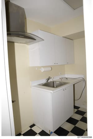 Photograph of a residential kitchen interior, likely from one of the properties under investigation. The image shows a compact kitchenette with white cabinetry, a stainless steel sink, modern range hood, and checkered black-and-white tile flooring. T