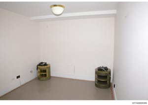 This is a photograph of an empty interior room, likely from an Epstein property during a federal investigation. The room shows white walls, a concrete floor with red base trim, a ceiling-mounted light fixture, and two cylindrical woven storage contai
