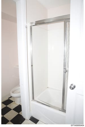This is a photograph of a bathroom interior showing a shower stall with glass sliding doors, white tile walls, a white toilet, and a black-and-white checkered tile floor. The image appears to be documentation of property conditions, likely from one o