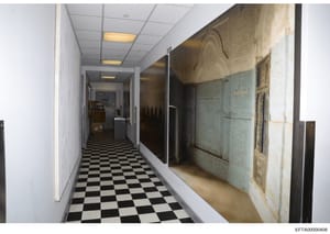 This photograph depicts a long interior hallway of what appears to be one of Epstein's properties, showing a distinctive black-and-white checkered floor, recessed ceiling lights, white walls, and ornate decorative panels or artwork on the right wall.
