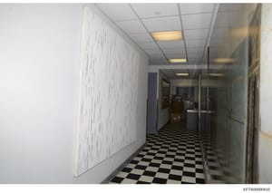 This photograph shows an interior hallway or corridor in what appears to be an office or institutional building. The hallway features a black-and-white checkered floor, fluorescent ceiling lighting, and glass partitions on the right side. On the left