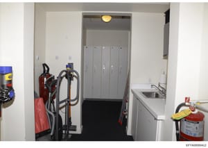 This is a photograph of an interior hallway or utility corridor showing maintenance and safety equipment including fire extinguishers, cleaning equipment, and an elevator. The space appears to be in a commercial or residential building. The image is 