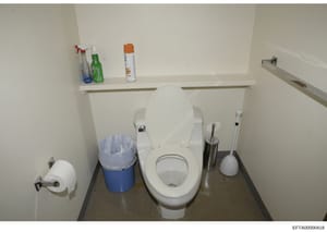 This is a photograph of a bathroom interior showing a white toilet, wall-mounted shelving with cleaning and personal care products, toilet paper holder, and waste bin. The image appears to be part of property documentation or evidence photography, wi