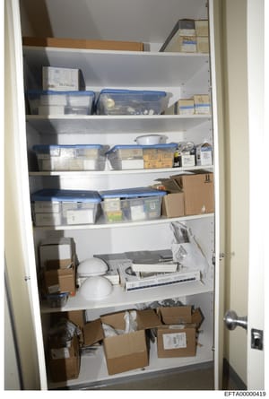 This photograph shows the interior of a storage closet containing multiple shelves stocked with blue plastic storage containers, cardboard boxes, office equipment (including what appears to be printers or scanners), and miscellaneous household items.