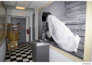 This is a photograph of an interior room, likely part of a property associated with the investigation. The image shows a bedroom or sleeping area with a bed frame that appears to be covered with white cloth or sheeting. The room contains basic furnis