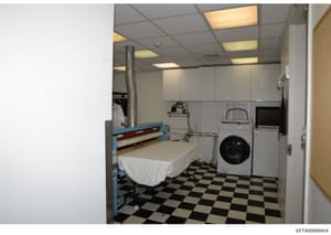 This photograph shows a combined bedroom and laundry area, likely part of staff quarters in one of Epstein's properties. The room contains a single bed with white linens, washing machine, dryer, and various household appliances. The space appears to 