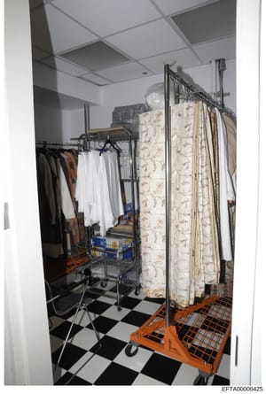This photograph shows a storage or dressing room containing multiple clothing racks with garments, storage containers, and household items. The room features a black and white checkered floor with orange plastic storage units, suspended ceiling tiles