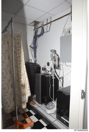 This is a photograph of an interior room space containing various electronic equipment, cables, and furnishings. The image shows a cluttered utility or technical space with black equipment boxes, overhead cable management, a patterned curtain, and ch