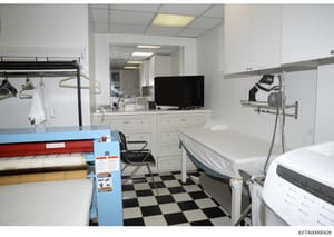 This photograph shows a commercial laundry room or housekeeping facility with professional-grade equipment and supplies. The space features a checkered floor, white cabinetry, rolling storage racks with garments and linens, a work desk with computer 