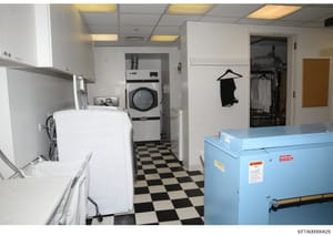 This photograph shows a commercial or institutional laundry room equipped with industrial-grade washing and drying equipment, including a white washing machine, a commercial dryer, and a large blue industrial washing or processing machine. The space 