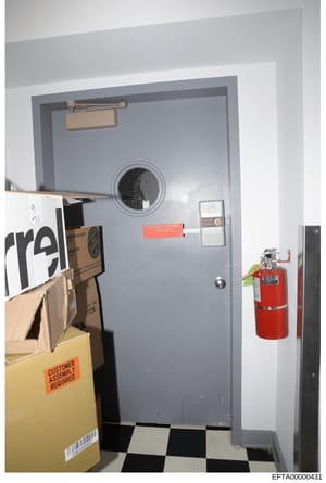 This is an evidence photograph showing a gray institutional door with security features, including a door closer, intercom system, and fire extinguisher. The door appears to be part of a secure facility or restricted area, with a black-and-white chec