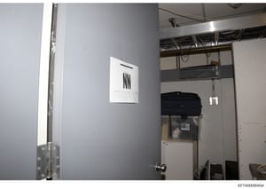 This is a photograph of an interior room, likely taken during the investigation, showing a closet or storage area with gray walls, exposed ceiling infrastructure, and stored items including a black bag/container. An evidence marker with tally marks i