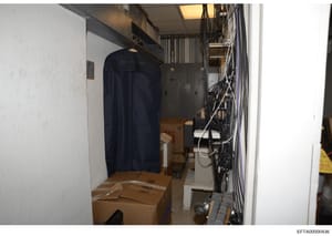 This is a photograph of a narrow storage space or utility closet containing various items including cardboard boxes, a garment bag with what appears to be designer branding (Ralph Lauren visible), electrical equipment, cable bundles, and other househ