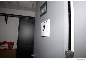 This is an evidence photograph showing the interior of a room or office space, featuring a black door, wall-mounted control system or alarm panel displaying circular indicators, shelving with a red 'FreshDirect' branded item, and various technical eq