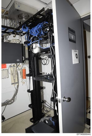 This photograph shows the interior of a network server closet or equipment cabinet containing various networking and IT infrastructure components. The image displays organized cabling, network switches, power distribution equipment, and cooling syste