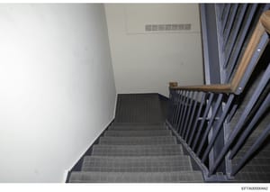 This is a photograph of an interior stairwell in what appears to be a residential or commercial building. The image shows a flight of stairs with dark carpeting, white walls, a wooden handrail, and HVAC venting visible at the top. The photo appears t