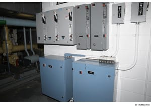 This photograph documents an electrical utility room containing multiple gray electrical panels mounted on a wall and blue transformer/distribution units positioned below. The image appears to be an evidence photo from a property inspection, showing 