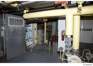 This photograph documents the basement mechanical room of a property, showing the building's HVAC, plumbing, and utility infrastructure including heating systems, piping, fire suppression equipment, and electrical panels. The image appears to be evid