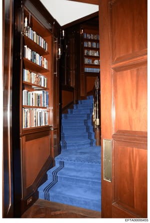 This is a photograph of an interior space featuring floor-to-ceiling wooden bookshelves with glass doors containing numerous books, dark wood paneling, and a blue patterned carpet or runner visible on stairs in the background. The image appears to be