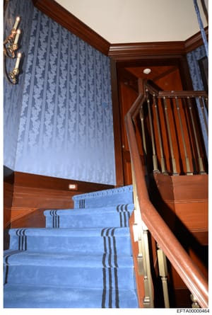This photograph documents the interior of a residential property, showing a wooden staircase with blue carpet runner and decorative striping. The image captures ornate wood detailing, blue damask-patterned wallpaper, brass wall sconces, and wood rail