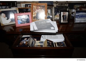 This is a photograph of a wooden desk displaying multiple framed photographs, loose photographs, and stacked documents. The image appears to be evidence documentation from the investigation, showing a collection of personal items and papers arranged 