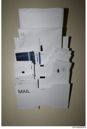 This is a photograph of collected mail items stored in what appears to be a wall-mounted organizer or evidence collection container labeled 'MAIL'. The items are heavily redacted with black bars obscuring addresses, names, and content details. This a