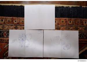 This photograph documents three sheets of white paper containing handwritten sketches and abstract drawings. The papers are photographed flat against a decorative rug with dark and rust-colored patterns. One sheet is dated 7/1/23 and contains various