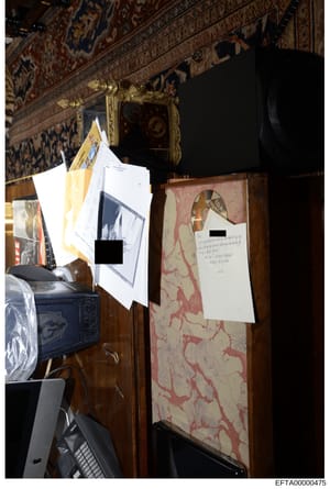 This is a photograph of an interior room showing luxurious décor including ornate ceiling with patterned panels, gilt-framed mirror, decorative furniture with red/cream patterned upholstery, and a blue ceramic piece. White documentation papers or evi