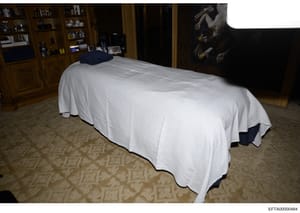 This is an evidence photograph showing a bedroom interior with a bed covered in white linens and a dark blue pillow, wooden cabinetry with decorative items, an ornate wooden door frame, patterned carpeting, and artwork on the wall. The photograph app