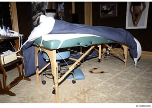 This photograph shows the interior of a bedroom containing a portable massage table with teal and blue cushioning, wooden frame, and blue draping. The room includes a wooden side table with personal items, artwork on the walls, and various furnishing