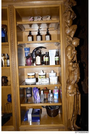 This photograph shows the contents of a wooden cabinet with glass doors containing various personal care items, cosmetics, fragrances, and grooming products. The image appears to be evidence documentation from a property search or inventory, showing 
