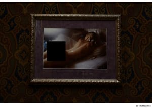 This is an evidence photograph showing a framed picture hung on a wall with distinctive ornate patterned wallpaper in gold and dark tones. The photograph within the frame contains a large black redaction box obscuring the primary content. The image a
