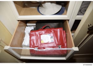 This is an evidence photograph showing a red leather briefcase with a document holder stored in a bedroom dresser drawer, alongside white clothing items and other personal effects. The briefcase appears to be seized as evidence during an investigatio