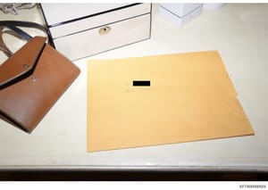 This is an evidence photograph showing a large manila envelope with a black redaction bar covering text, alongside a brown leather shoulder bag and white storage boxes on what appears to be a desk or work surface. The envelope contains handwritten te