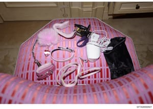 This is an evidence photograph showing various personal items and accessories arranged on pink striped bedding in what appears to be a residential bedroom. The items include shoes, electronic devices, jewelry, and accessories. The photo was taken as 