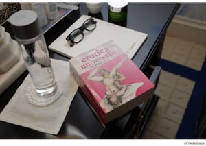 This is an evidence photograph showing a pink-covered book titled 'Erotica Universalis' (published by Taschen) displayed on a nightstand or table alongside personal items including eyeglasses, a water bottle, toiletries, and a green bottle. The photo