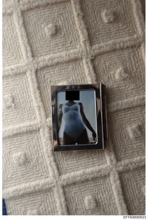 This is a photograph of a framed image placed on a cream-colored quilted or textured fabric surface. The image within the frame shows a figure in a light blue/gray garment with the face redacted by a black rectangle. The photo appears to be evidence 