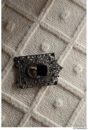 This photograph shows a decorative metal-framed object with ornate filigree work and blue stone or crystal embellishments, positioned on a cream-colored quilted fabric surface. The center contains a mirrored or reflective rectangular opening with a r