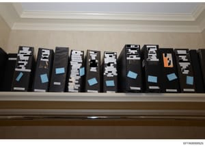 This photograph documents a shelf displaying approximately 10-11 black archival storage boxes containing investigation case files, evidence, and documents related to the Epstein case. The boxes are labeled with evidence tags and blue index stickers i