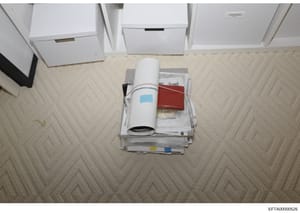 This is an overhead photograph of evidence collected during the investigation, showing a stack of documents, a rolled architectural or technical drawing with a blue label, a red leather-bound book or journal, and various papers arranged on a beige ca