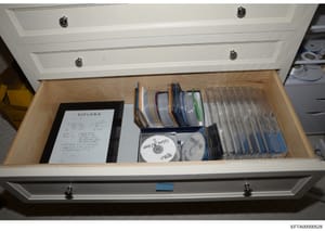This photograph shows the contents of a white drawer seized during the investigation, containing a framed diploma document, numerous CDs organized in storage cases, and loose media. The items appear to be personal documents and digital materials stor