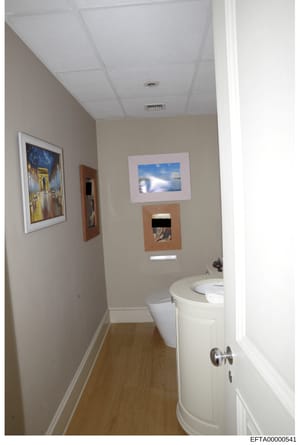 This is an evidence photograph depicting the interior of a bathroom, showing a toilet, sink vanity, and several framed artworks on the walls. The photo documents the interior condition and furnishings of a property relevant to the investigation. The 