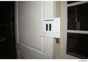 This is a photograph of an evidence item mounted on a white wall between two windows, displaying what appears to be two dark rectangular objects affixed to a white documentation board with a reference label and information grid. The photo was taken a