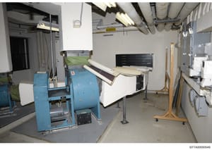 This is a photograph of an underground or basement utility space containing industrial laundry and HVAC equipment. The room shows blue commercial washing machines, white ductwork, a black tool chest, rolled materials, and various utility connections.