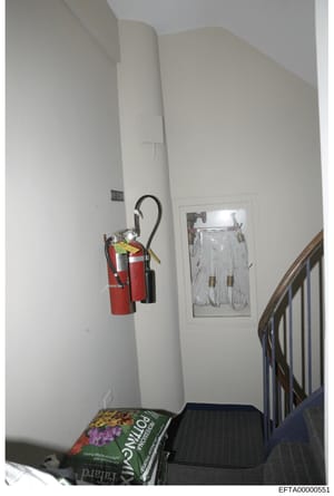 This is an evidence photograph showing an interior stairwell area, likely from one of Epstein's properties. The image captures fire safety equipment (two red fire extinguishers), electrical conduit, framed artwork on the wall, a wooden stair rail, an