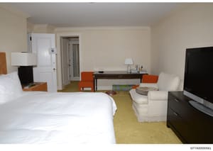 This photograph shows a hotel suite interior featuring a bedroom with a queen-sized bed, a living area with upholstered seating, and a work desk. The image appears to be documentation of accommodations, possibly related to properties or residences un