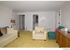 This is a photograph of a residential living room interior, showing furnished seating areas, carpeting, and doorways leading to adjacent rooms. The image appears to be documentation of a property space, likely part of an evidence or asset inventory r