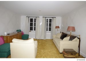 This is a photograph of a residential interior room showing furnishings and décor. The room contains multiple cream-colored seating pieces, green accent furniture, wooden dressers, and dual sets of French doors with cream curtains. The space appears 